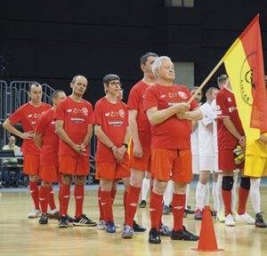 Piłkarze w czerwonych strojach, stoją jeden za drugim, pierwszy trzyma flagę czerono żółtą